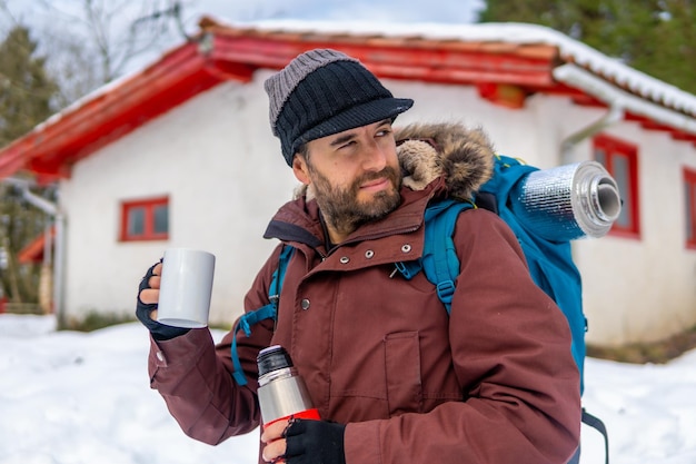 Homem bebendo café de uma garrafa térmica quente no inverno na neve ao lado de uma cabana depois de dormir nela