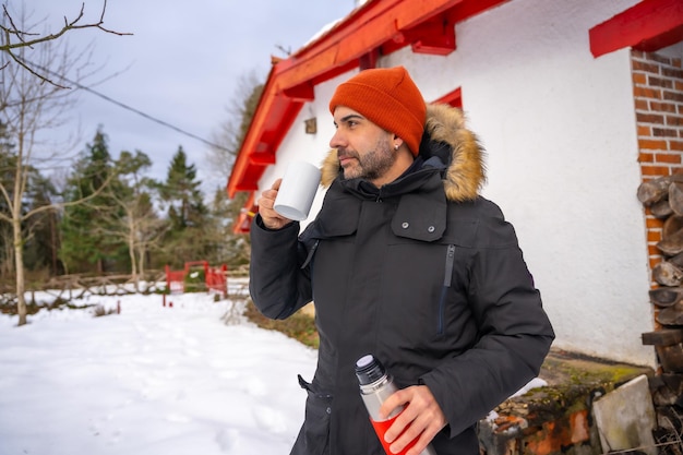 Homem bebendo café de uma garrafa térmica quente no inverno na neve perto da madeira da lareira