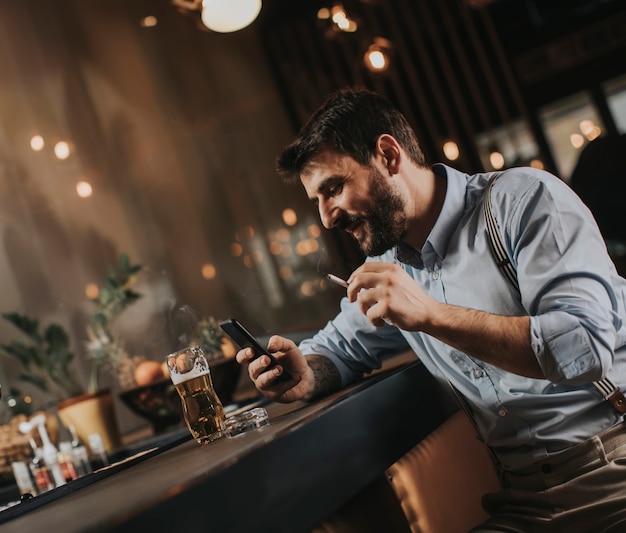 Homem bebendo cerveja, fumando cigarro e olhando para o celular no pub