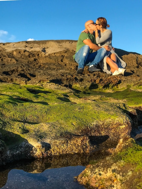 Homem beijando mulher enquanto está sentado em uma formação rochosa contra o céu azul