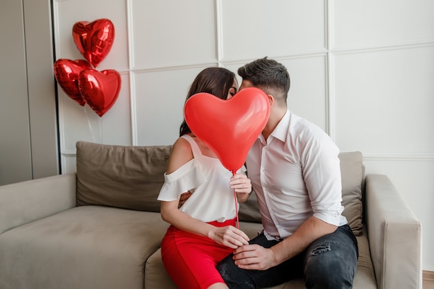 Homem beijando mulher por trás da forma do coração com balões vermelhos em casa sentado no sofá