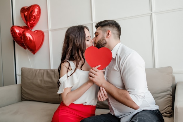 Foto homem beijando mulher por trás da forma do coração com balões vermelhos em casa sentado no sofá