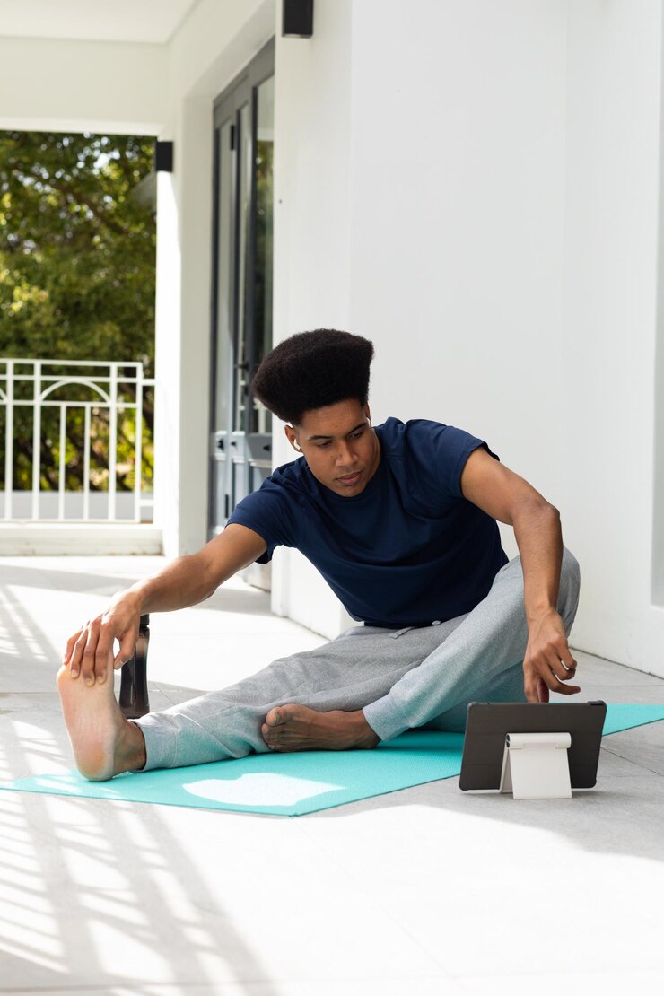 Foto homem biracial a fazer ioga, alongamento e usar tablet sentado na varanda ensolarada, espaço de cópia. bem-estar, fitness, comunicação e estilo de vida saudável, inalterado.