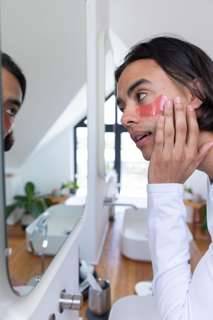 Homem biracial feliz a olhar-se no espelho, a aplicar manchas nos olhos na casa de banho, a passar tempo de qualidade em casa sozinho, cuidados com a pele, vida doméstica e conceito de estilo de vida.