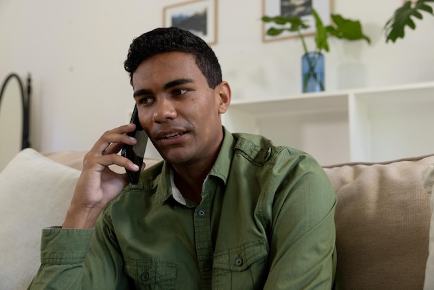 Homem biracial feliz sentado no sofá falando em smartphone em casa