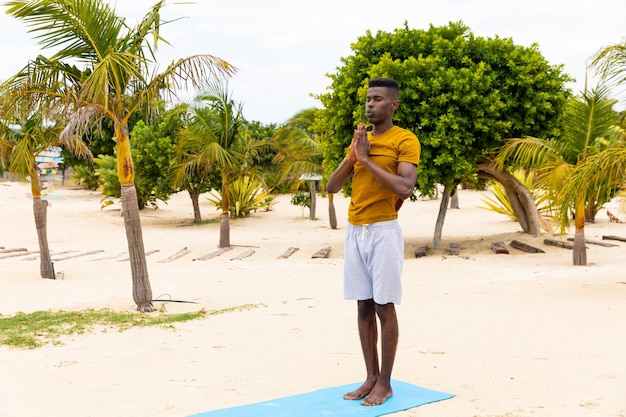 Homem birracial com os olhos fechados praticando ioga meditação em pé na praia ensolarada, inalterado. Estilo de vida saudável, bem-estar, relaxamento, verão e férias.