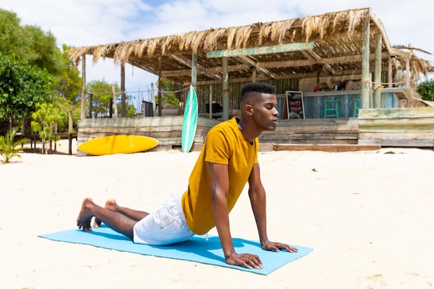 Homem birracial focado praticando ioga, estendendo-se no tapete na praia ensolarada, inalterado. Pose de cobra, estilo de vida saudável, bem-estar, verão e férias.