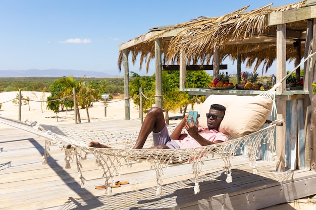 Homem birracial relaxado em óculos de sol, deitado na rede usando smartphone no ensolarado bar da praia. Relaxamento, comunicação, estilo de vida, tempo livre, verão e férias, inalterados.