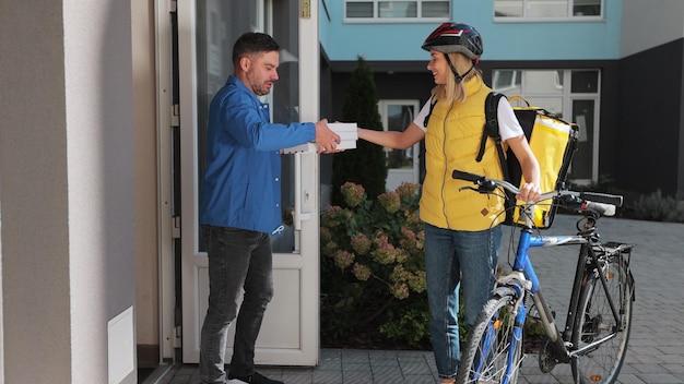 Homem bonito abre a porta para uma entregadora de pizza usando mochila térmica amarela em uma bicicleta e recebe caixas de papelão de pizza Conceito de entrega de comida