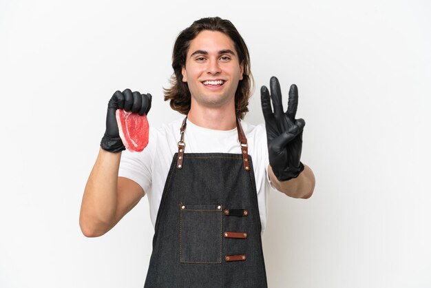 Homem bonito açougueiro vestindo um avental e servindo carne fresca isolada no fundo branco feliz e contando três com os dedos