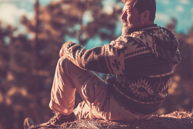 Foto homem bonito adulto aproveite o sol no rosto e relaxe, sente-se no chão com árvores e bosques ao fundo - pessoas felizes e livres ao ar livre - conceito de viagens e turismo de fim de semana