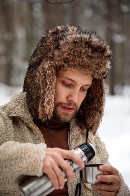 Homem bonito alpinista em roupas quentes derrama chá em caneca de garrafa térmica na floresta de inverno Conceito de temporada Foco seletivo