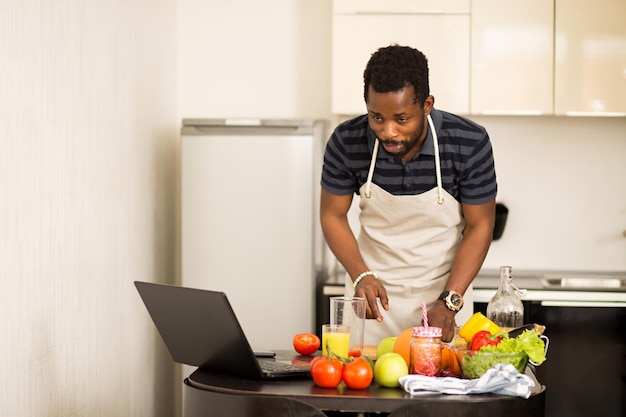 Homem bonito americano africano vestindo camiseta listrada, avental, procurando receita no computador laptop e cozinhando vegetais na cozinha leve em casa. Alimentação saudável, dieta, conceito de tecnologia.