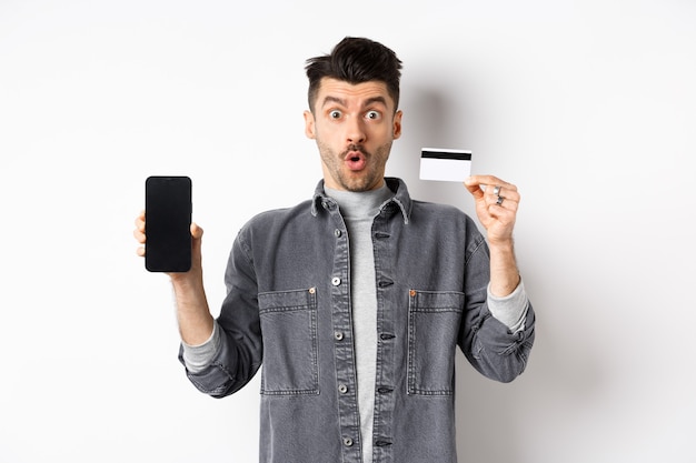 Homem bonito animado mostrando o cartão de crédito de plástico e a tela do telefone móvel, de pé sobre um fundo branco.