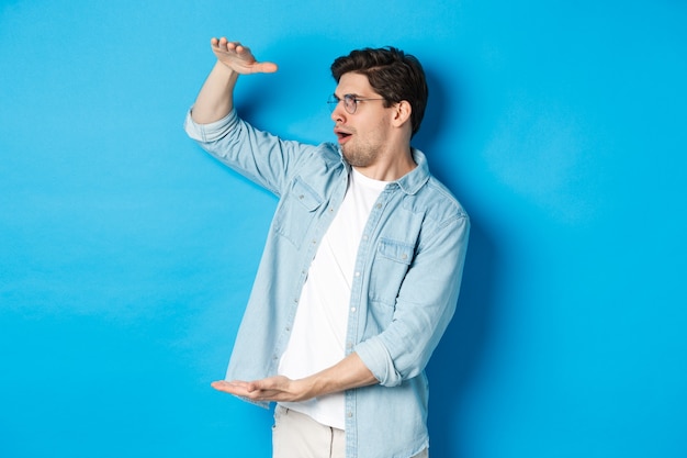 Homem bonito animado mostrando um objeto de grande tamanho e parecendo surpreso, de pé sobre a parede azul