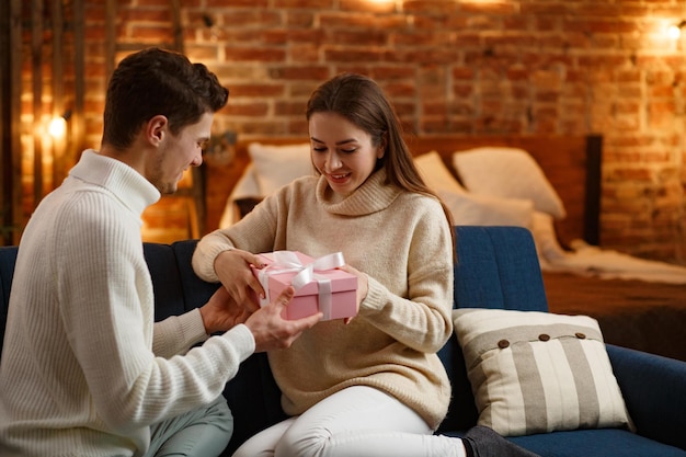 Homem bonito, apresentando um presente para sua linda garota e sorrindo. Lindo casal jovem em casa desfrutando de passar algum tempo juntos. Férias de inverno, celebrações do Natal, conceito de ano novo.