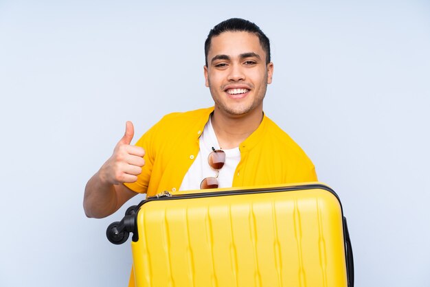 Homem bonito asiático isolado na parede azul de férias com mala de viagem e polegar para cima