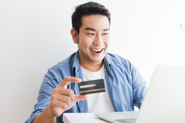 Homem bonito asiático na camisa azul usando cartão de crédito com o computador portátil, compras on-line