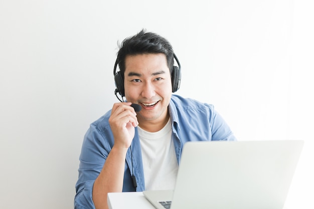 Homem bonito asiático na camisa azul usando laptop com fone de ouvido falando sorriso e cara feliz
