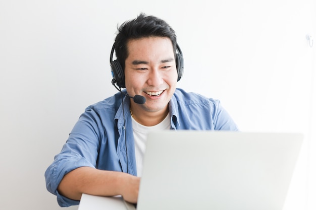 Homem bonito asiático na camisa azul usando laptop com fone de ouvido falando sorriso e cara feliz