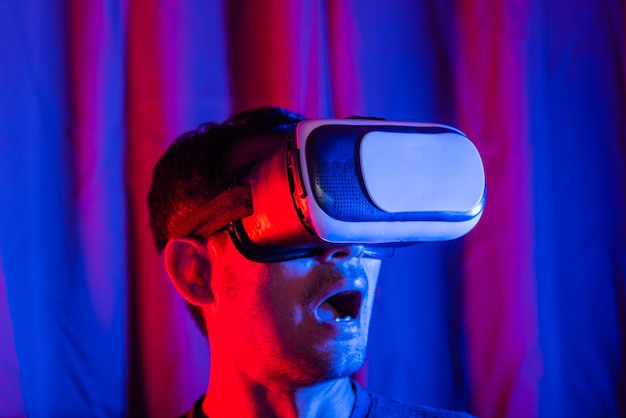 Homem bonito asiático usa capacete VR animado emocionalmente em casa fundo roxo e azul