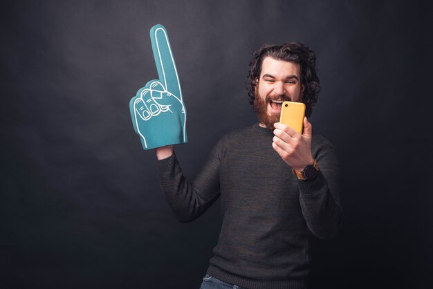 Homem bonito barbudo animado apontando com uma luva em leque e olhando para um smartphone amarelo