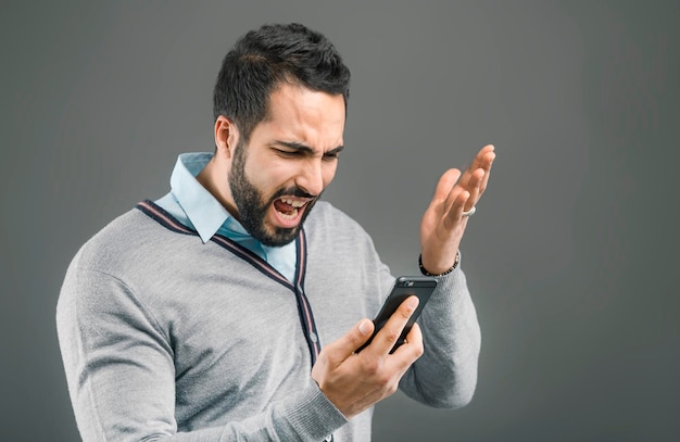 Homem bonito barbudo com expressão de cara feia, segurando o smartphone na mão em fundo cinza isolado