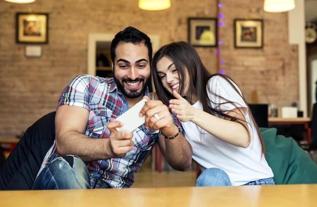 Homem bonito barbudo e menina morena se divertindo jogando jogo online no smartphone no conceito de emoções felizes de quarto elegante