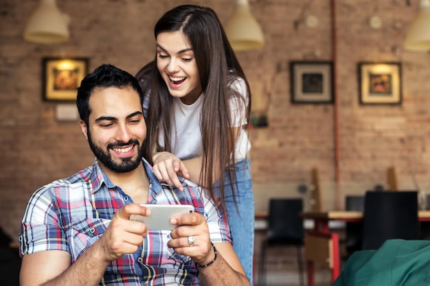 Homem bonito barbudo e menina morena se divertindo jogando jogo online no smartphone no conceito de emoções felizes de quarto elegante