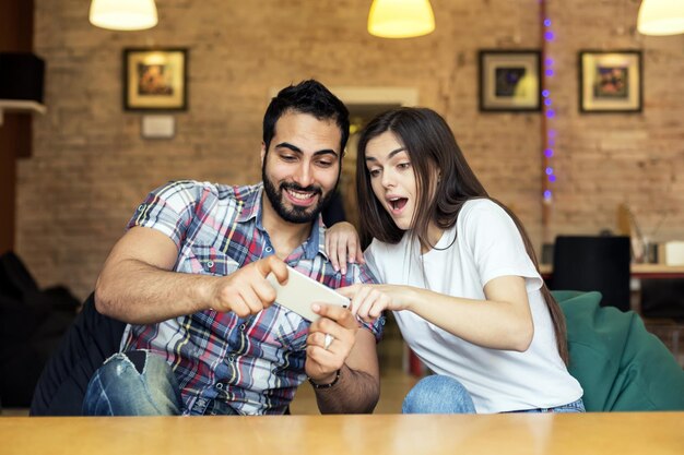 Homem bonito barbudo emocional e menina morena se divertindo jogando jogo online no smartphone em um quarto elegante