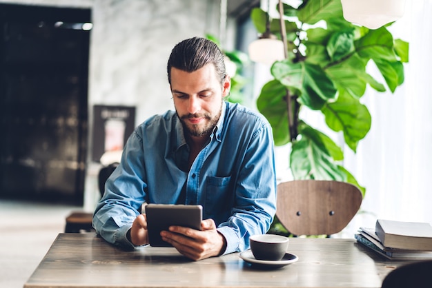 Homem bonito barbudo hipster relaxante usando computador tablet com café enquanto está sentado à mesa no café. Conceito de tecnologia e comunicação