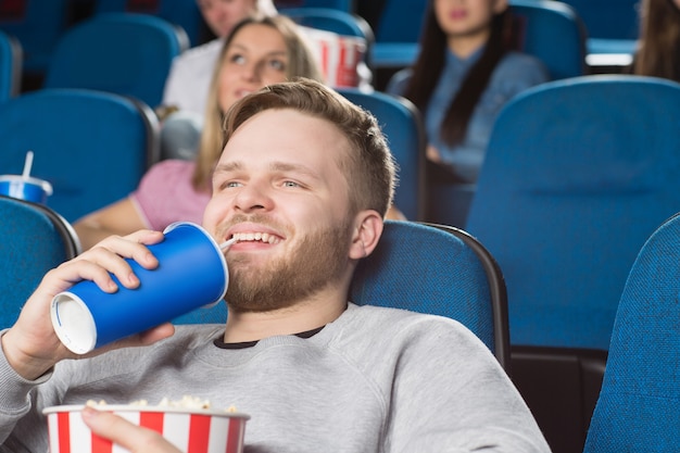 homem bonito bebendo segurando um balde cheio de pipoca sorrindo alegremente assistindo filmes no cinema