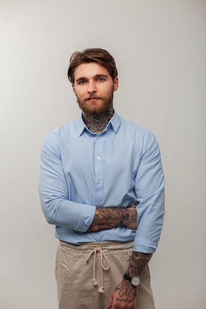 Homem bonito brutal hipster com penteado de barba e tatuagens na moda camisa azul fica em um fundo cinza