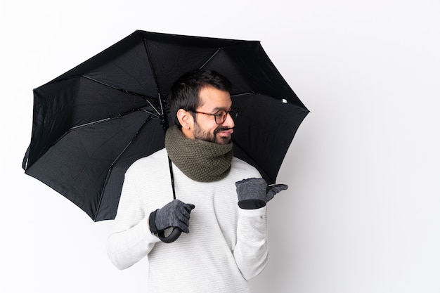 Homem bonito caucasiano com barba segurando um guarda-chuva sobre parede branca infeliz e apontando para o lado