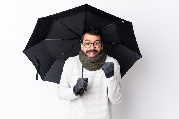 Homem bonito caucasiano com barba segurando um guarda-chuva sobre parede branca isolada, comemorando uma vitória na posição de vencedor