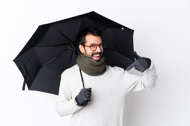 Homem bonito caucasiano com barba segurando um guarda-chuva sobre parede branca isolada, orgulhoso e satisfeito