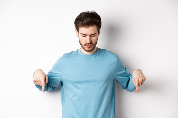 Homem bonito caucasiano com barba, vestindo camisa azul, olhando e apontando para baixo no banner de logotipo interessante, de pé sobre fundo branco.