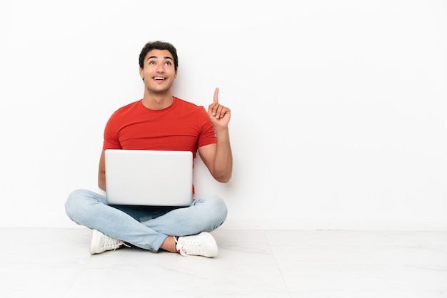 Homem bonito caucasiano com um laptop sentado no chão apontando para cima e surpreso