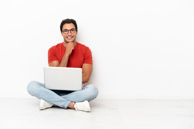 Homem bonito caucasiano com um laptop sentado no chão com óculos e sorrindo