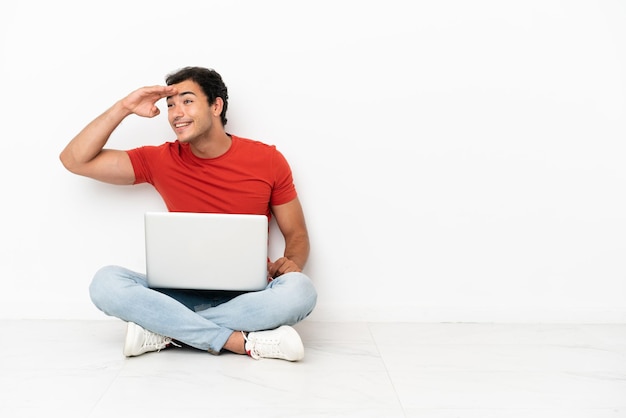 Homem bonito caucasiano com um laptop sentado no chão olhando para longe com a mão para olhar algo