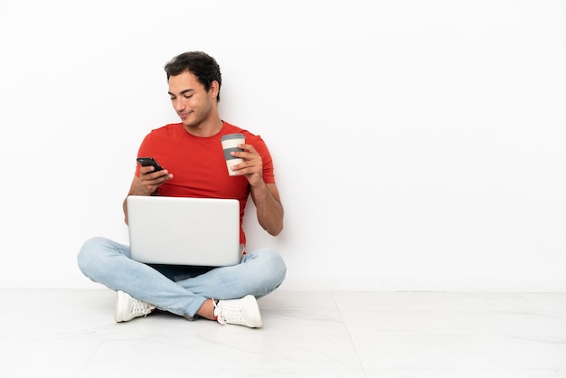Homem bonito caucasiano com um laptop sentado no chão segurando um café para levar e um celular