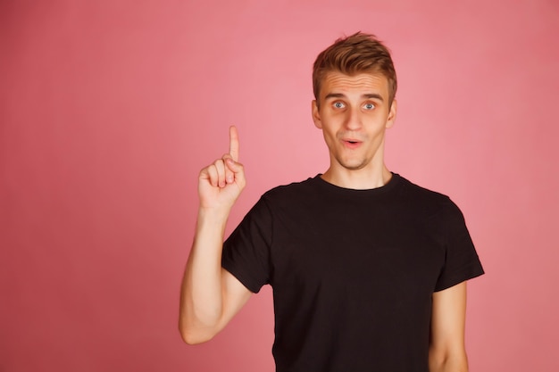 Homem bonito caucasiano em camiseta preta oferece uma ideia, mostrando seu dedo indicador isolado no fundo rosa. Retrato de cara perplexo. Conceito de percepção repentina. Espaço de direitos autorais para o site