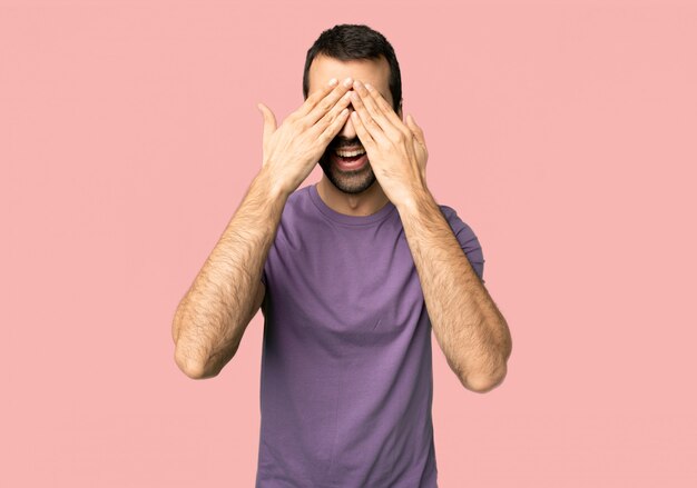 Foto homem bonito, cobrindo os olhos pelas mãos. surpreso para ver o que está à frente no fundo rosa isolado