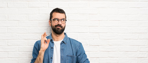 Homem bonito com barba ao longo da parede de tijolos brancos com dedos cruzando e desejando o melhor