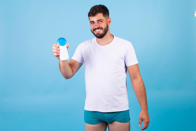 Homem bonito com barba de férias vestindo trajes de banho segurando o frasco de loção protetor solar parecendo positivo e feliz em pé e sorrindo com um sorriso confiante mostrando os dentes