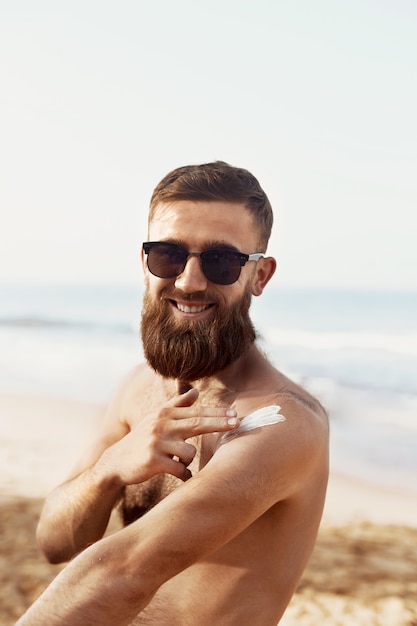 Homem bonito com barba, em óculos de sol, tomando banho de sol com loção de protetor solar no corpo no verão. Modelo de fitness masculino bronzeamento artificial usando creme de bloco solar para bronzeamento saudável.