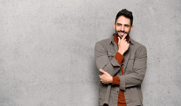 Homem bonito, com barba, olhando para o lado com a mão no queixo sobre a parede texturizada