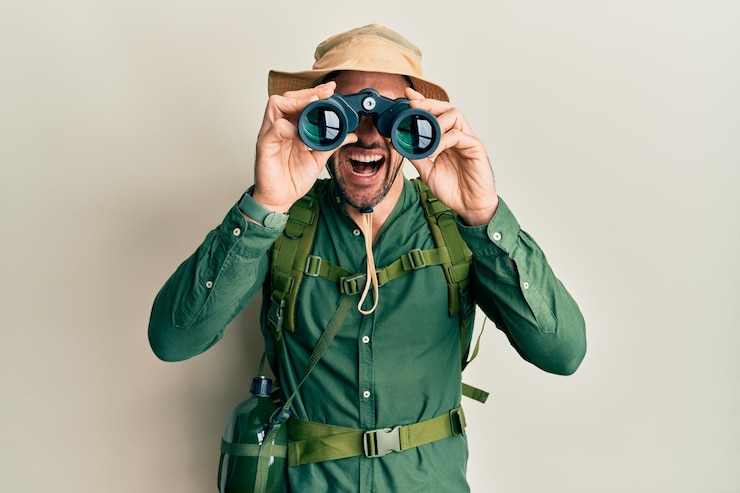 Foto homem bonito com barba usando chapéu de explorador olhando através de binóculos sorrindo e rindo alto porque uma piada engraçada e maluca