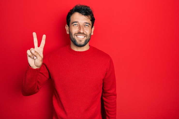 Foto homem bonito com barba vestindo suéter vermelho casual sorrindo olhando para a câmera mostrando os dedos fazendo o sinal de vitória número dois