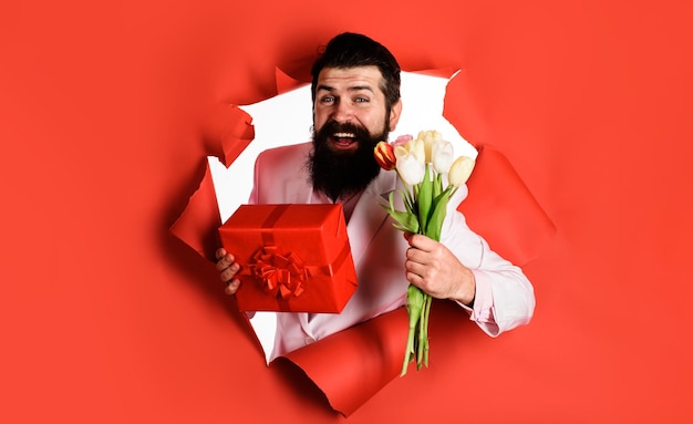 Homem bonito com caixa de presente e buquê de flores empresário com presente e tulipas olhando através